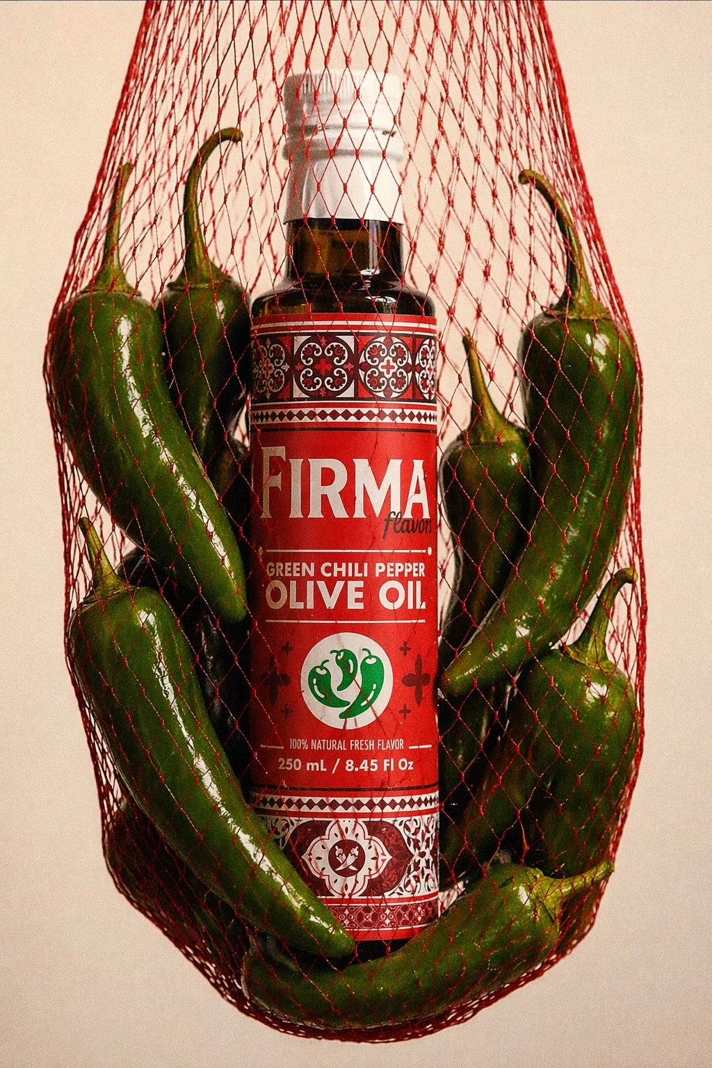 A bottle of Firma Green Chili Pepper Olive Oil nestled inside a red mesh bag filled with green chili peppers, set against a plain beige background.