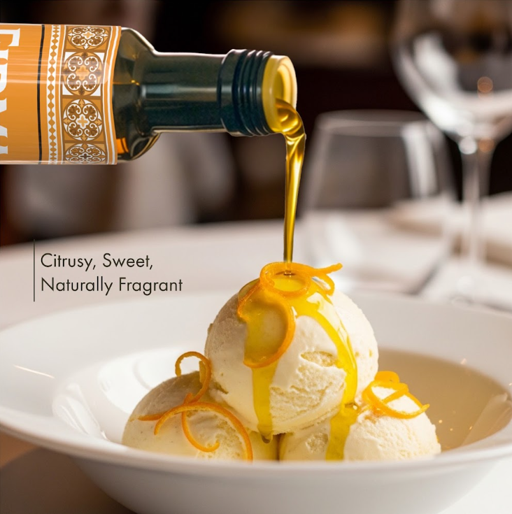 A bottle of orange-flavored olive oil is pouring a golden stream over three scoops of vanilla ice cream in a white bowl, with orange zest drizzled on top. Text on the left reads 'Citrusy, Sweet, Naturally Fragrant'.